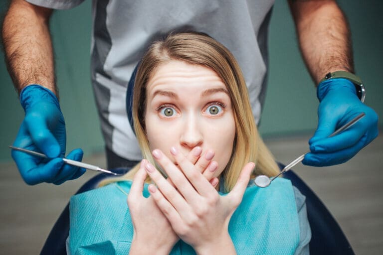 A young woman in a dental chair covers her mouth with both hands, looking surprised or scared, while a dentist wearing blue gloves holds dental tools near her face.