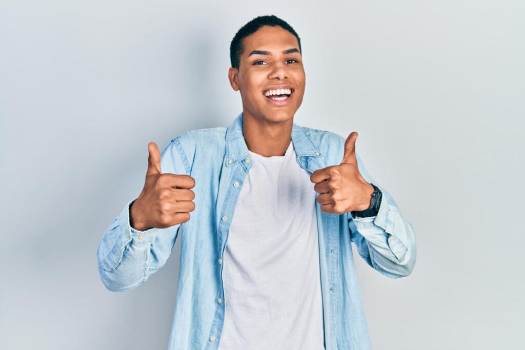A young person smiles broadly while giving two thumbs up, wearing a light blue denim shirt over a white t-shirt and standing against a plain light background—perfect for showcasing their happy visit to the Northville dentist.