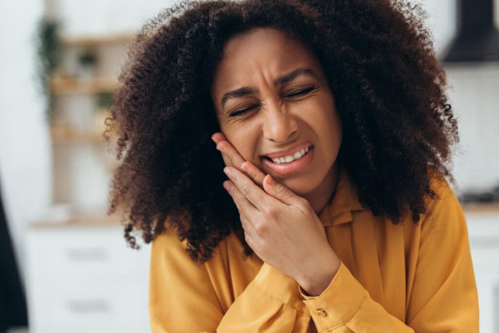 A woman with curly hair wearing a yellow shirt holds her cheek and winces in pain, suggesting she has a toothache or jaw pain, in a bright indoor setting.