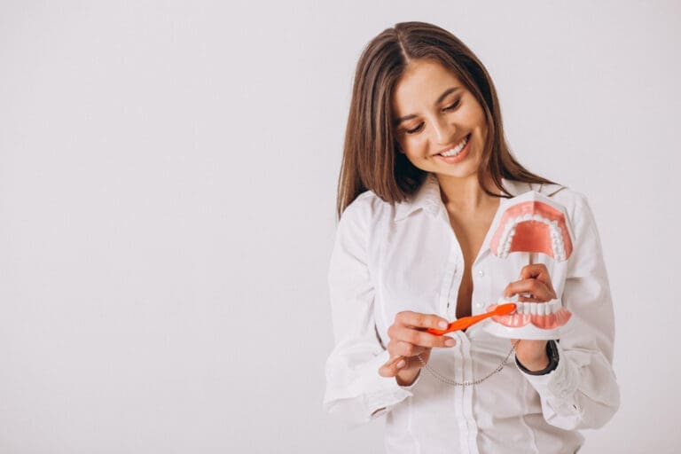 A woman in a white shirt smiles while holding a large model of teeth and demonstrating brushing with an oversized toothbrush against a plain white background.