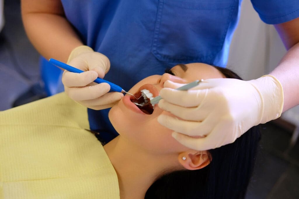 A dentist wearing gloves examines a woman’s open mouth with dental tools while she lies back in a dental chair, preparing for a dental procedure.