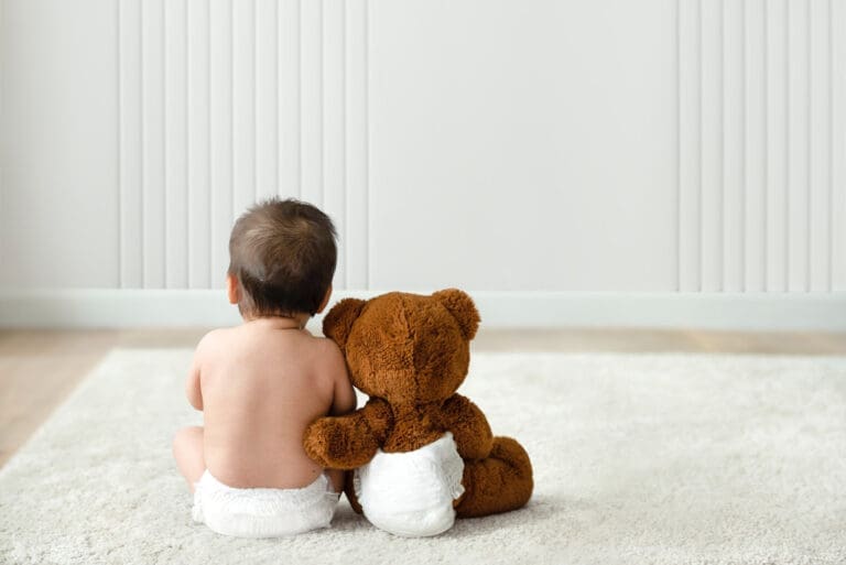 A baby wearing a diaper sits on a soft white rug, facing away from the camera, next to a brown teddy bear that is also wearing a diaper. Both are positioned close together in a minimalistic room.