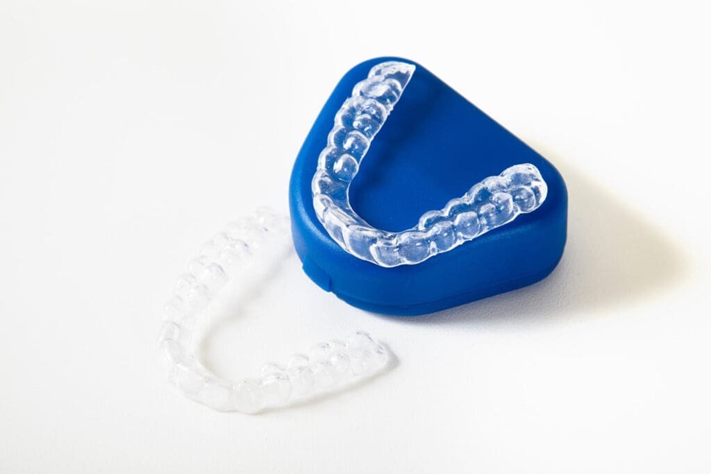 A pair of clear dental aligners, one resting inside a blue storage case and the other lying on a white surface next to it.