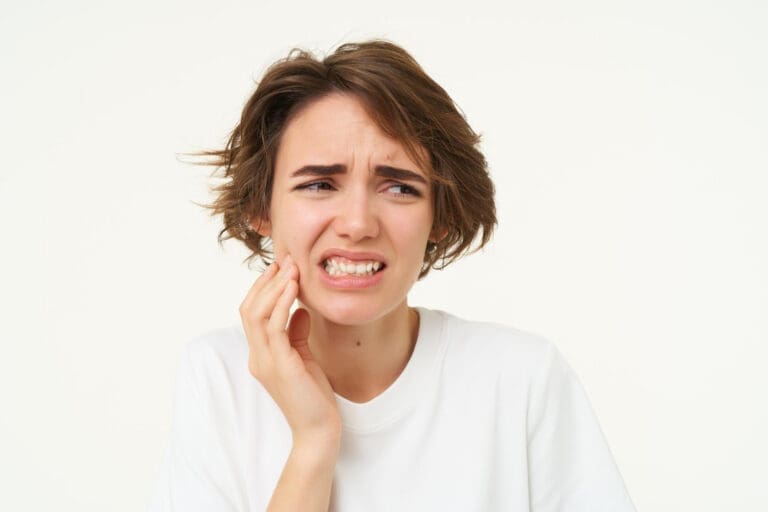 A young person with short brown hair in a white shirt touches their cheek and grimaces in discomfort, possibly due to tooth or jaw pain, against a plain white background.