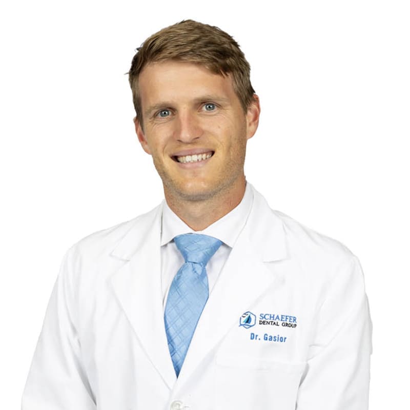 A smiling man with light brown hair wears a white dental coat, light blue tie, and a white shirt. "Schaefer Dental Group" and "Dr. Gasior" are embroidered on the coat, representing a trusted Northville dentist. The background is white.