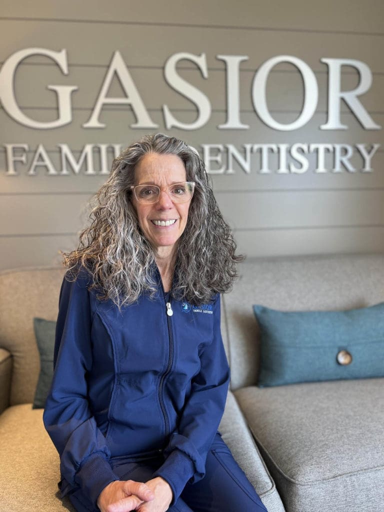 A woman with long curly gray hair and glasses, wearing a navy blue uniform, sits on a couch in front of a wall sign that reads GASIOR FAMILY DENTISTRY at this Northville dental office.