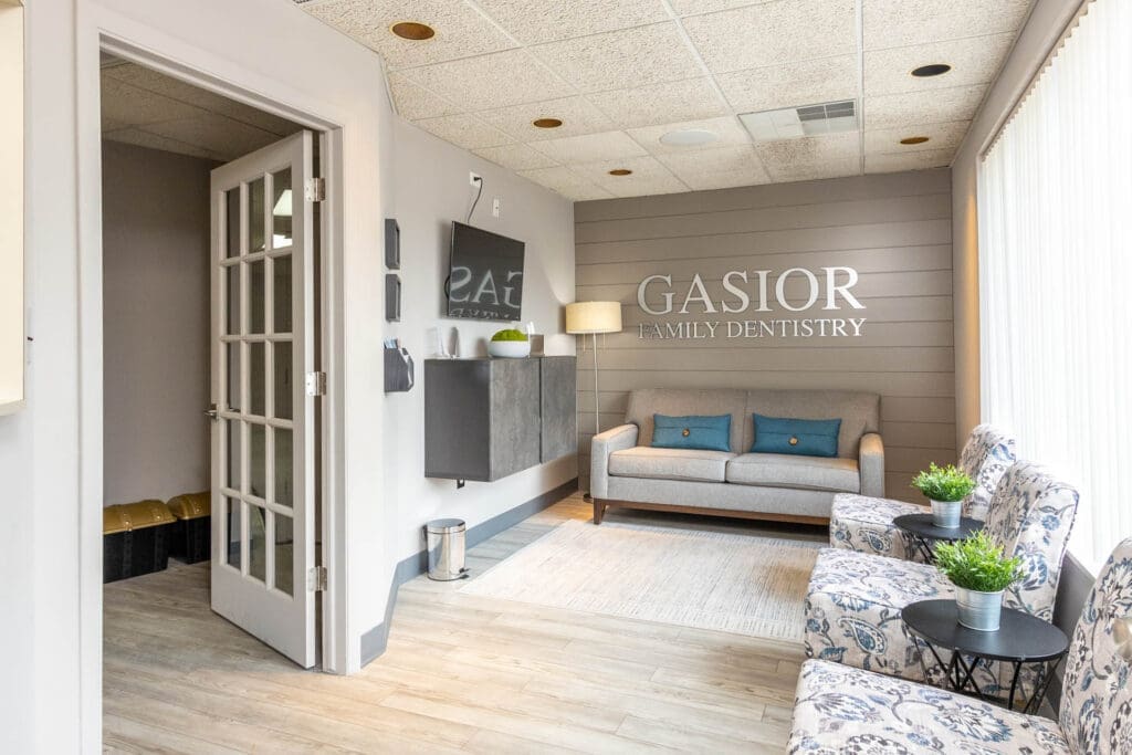 A clean, modern Northville dental office waiting room features a gray couch, two patterned chairs, potted plants, a rug, and a wall sign reading GASIOR FAMILY DENTISTRY. Sunlight streams through tall windows with blinds.
