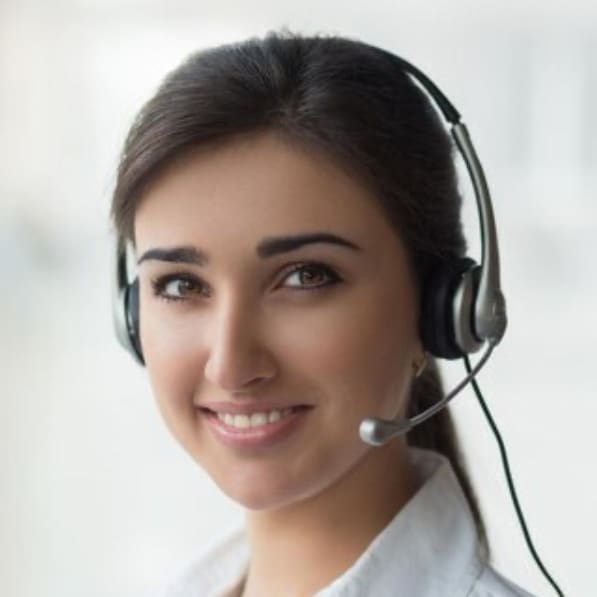 A woman with long dark hair, wearing a white top and a headset with microphone, smiles while looking at the camera. The softly blurred background suggests she could be assisting patients at a Northville dental office.