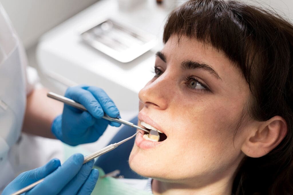 A woman sits in a dental chair with her mouth open while a Northville dentist wearing blue gloves examines her teeth using dental tools.