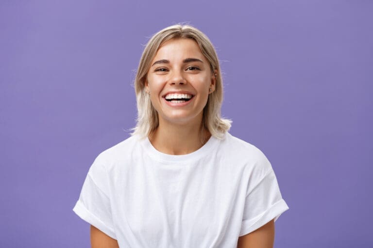 A young person with short blond hair wearing a white t-shirt is smiling widely while standing in front of a plain purple background, showing off their bright smile after visiting a Northville dentist.