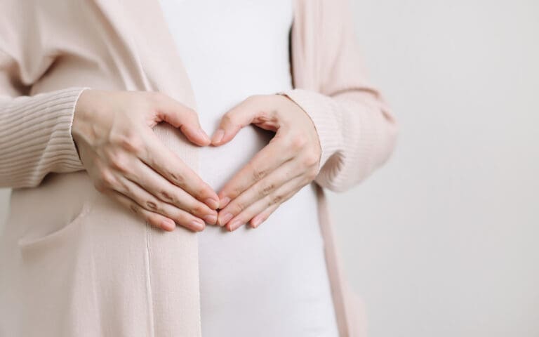 A pregnant woman in a light pink cardigan forms a heart shape with her hands over her belly against a plain, light background, highlighting the warmth often found at a Northville dental office.
