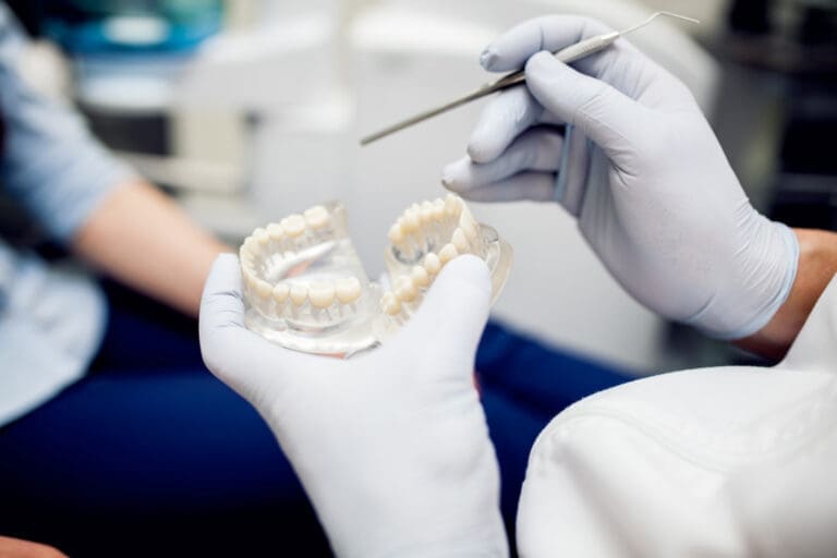 A Northville dentist wearing white gloves holds a dental model and uses a dental tool while discussing with a patient in the clinic.