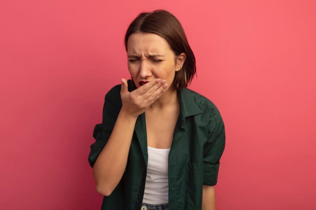 A woman with short brown hair in a green shirt and white top stands against a pink background, covering her mouth with her hand and looking nauseous—perhaps reminding you to visit your Northville dentist.